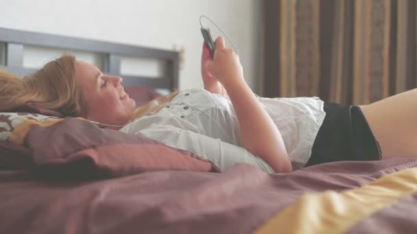 Beautiful young girl lying on the bed and use smartphone smiling - Felvétel, videó