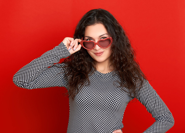 schönes Mädchenporträt posiert und flirtet auf rotem Hintergrund, lange lockige Haare, Sonnenbrille in Herzform, Glamour-Konzept - Foto, Bild