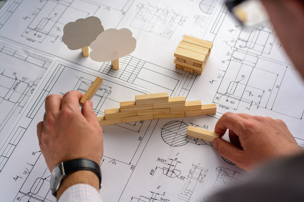 Man architect draws a plan, graph, design, geometric shapes by pencil on large sheet of paper at office desk and builds model house from wooden blocks (bars) - Photo, Image