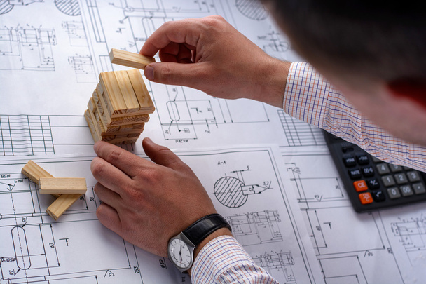 Man architect draws a plan, graph, design, geometric shapes by pencil on large sheet of paper at office desk and builds model house from wooden blocks (bars) - Photo, Image