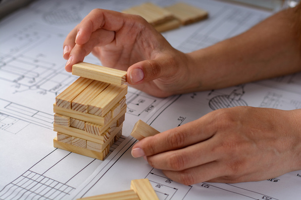 Man architect trekt een plan, ontwerp, grafiek en geometrische vormen door potlood op groot vel papier op een bureau en bouwt model huis van houten blokken (balken) - Foto, afbeelding