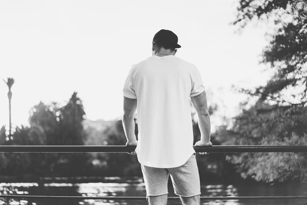 Bearded Muscular Man Wearing Blank t-shirt, snapback cap and shorts in summer time.Relaxing time near the lake.Green City Garden Park Sunset Background. Back view. Horizontal Mockup.Black White - Photo, image