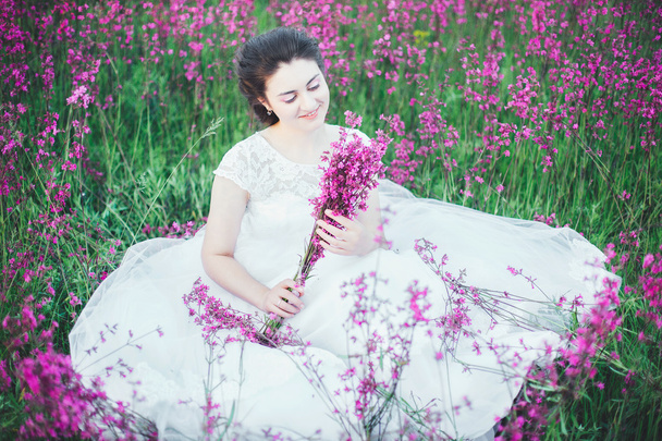 όμορφη νύφη σε ένα πεδίο λουλούδι. Το κορίτσι σε ένα λευκό φόρεμα με μια ανθοδέσμη σε ένα πεδίο καλοκαίρι στο ηλιοβασίλεμα - Φωτογραφία, εικόνα