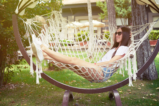 Young smiling woman in sunglasses resting in hammock in backyard - 写真・画像
