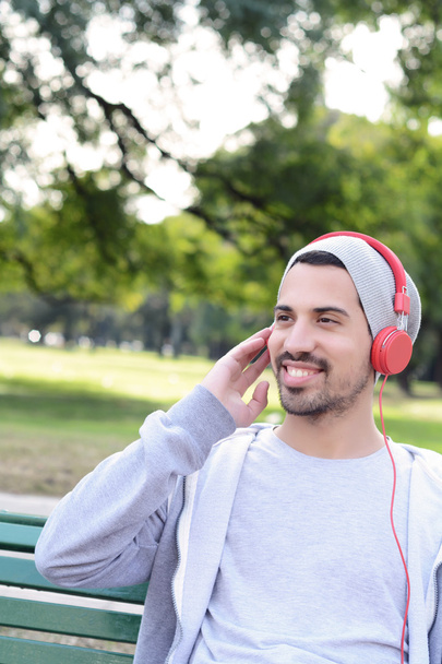 Νεαρός άνδρας, να ακούτε μουσική με ακουστικά που κάθεται στο πάρκο ben - Φωτογραφία, εικόνα
