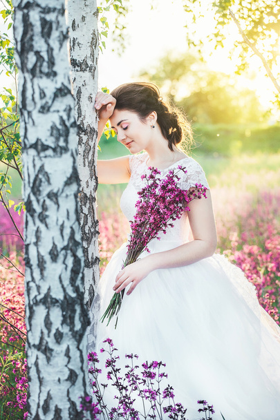 The girl in a white dress with a bouquet in a summer field at sunset stands near the birch - Fotó, kép