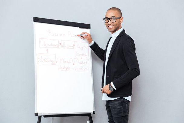 Smiling african man in glasses writing business plan on flipchart - 写真・画像