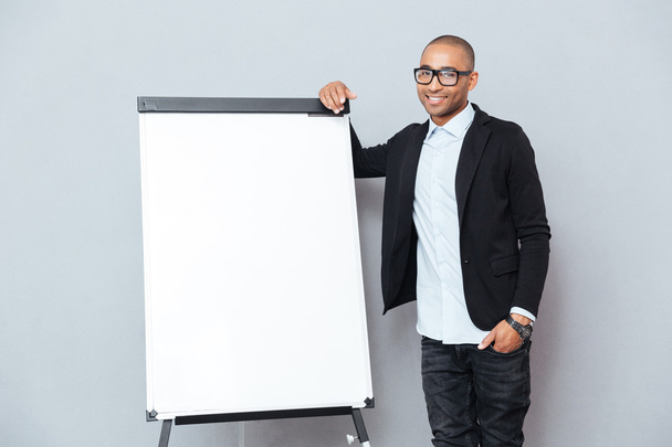 Cheerful african young businessman in glasses standing near blank flipchart - Fotó, kép