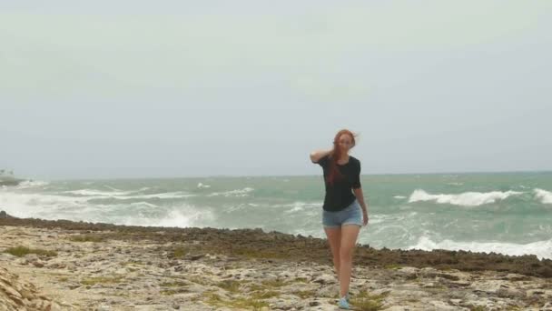 Mujer atractiva joven con el pelo rojo largo vestido en pantalones cortos caminando en la costa frente al mar de tormenta, gran ángulo, cámara lenta
 - Metraje, vídeo