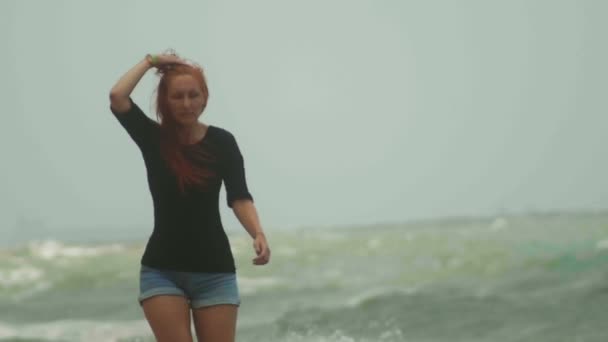 Mladá atraktivní žena s dlouhé splývavé červené vlasy vlasy oblečený v krátké šortky procházky podél moře bouří, zpomalené - Záběry, video