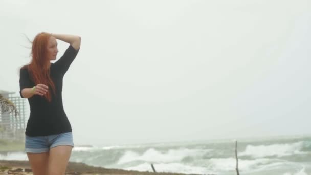 Młoda atrakcyjna kobieta z długimi rudymi włosami ubrany w skrócie szorty spaceru wzdłuż brzegu morza burza, zwolnionym tempie - Materiał filmowy, wideo