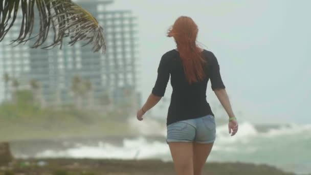 Jonge aantrekkelijke vrouw met lange rode haren gekleed in korte broek lopen aan de kust tegenover de bestorming zee, zet weg, slow-motion - Video