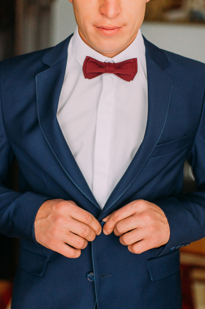 Man with burgundy bow tie and white shirt buttons up his dark blue blazer - Фото, изображение