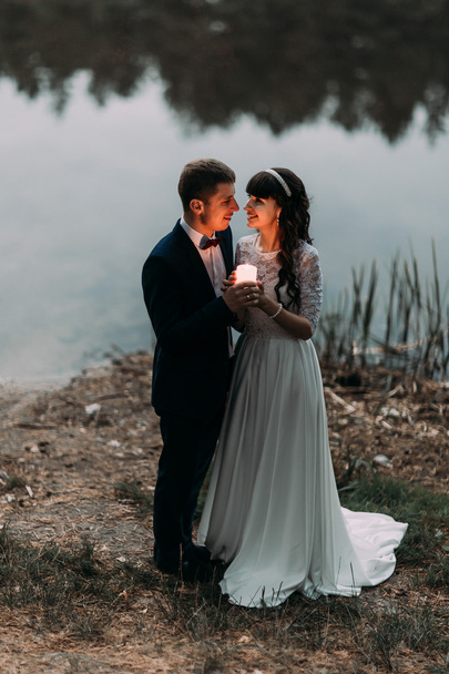 Groom and his charming new wife with burning candle on the shore of forest lake at evening - Foto, Imagen