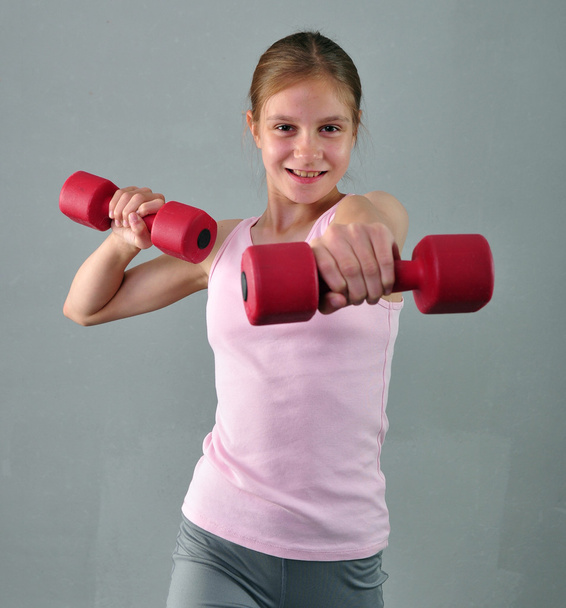 Teenage sportive girl is doing exercises to develop muscles on grey background. Sport healthy lifestyle concept. Sporty childhood. Teenager exercising with wieghts. - Fotó, kép