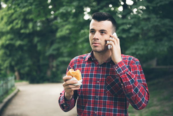Handsome young man eating sandwich autdoor. He is holding a phone - Fotografie, Obrázek