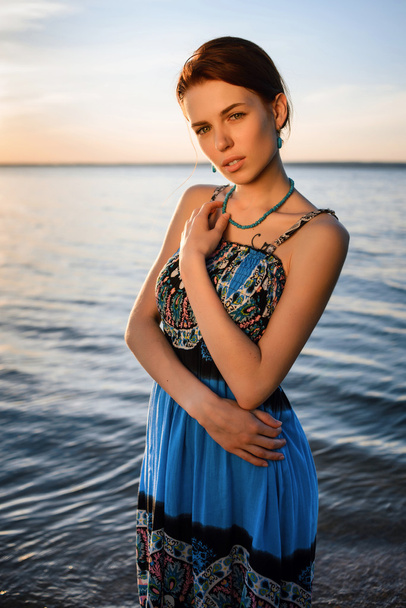 Young beautiful girl in blue dress posing at sea coast against blue sky at sunset - Photo, Image