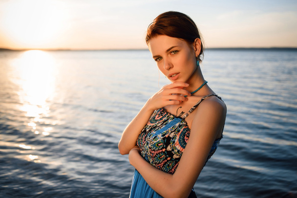 Young beautiful girl in blue dress posing at sea coast against blue sky at sunset - Zdjęcie, obraz