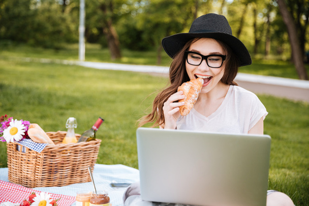 Happy woman eating croissant and using laptop outdoors - Fotoğraf, Görsel