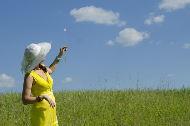 Το κορίτσι κρατά ένα λουλούδι σε ένα καπέλο και ένα κίτρινο φόρεμα μια πικραλίδα στο μπλε του ουρανού - Φωτογραφία, εικόνα