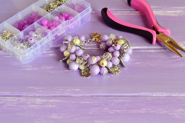 Beautiful bracelet with lilac and white plastic beads, metal flowers and leaves. Organizer with different beads, steel decorative pendants and rings. Pliers. Homemade jewelry concept - Photo, Image