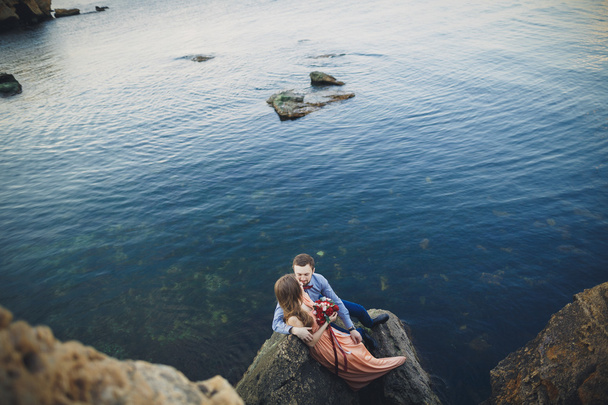 Wedding couple sitting on large stone around blue sea - Fotografie, Obrázek