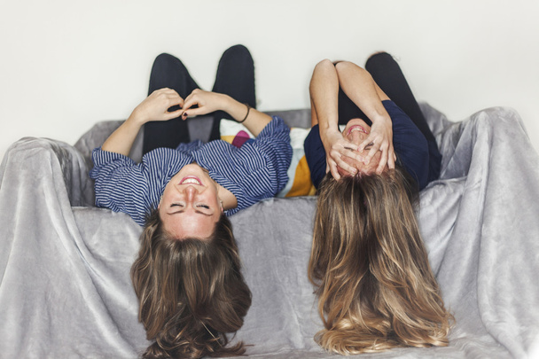 Two girls laying upside down on a grey sofa at home smiling - 写真・画像