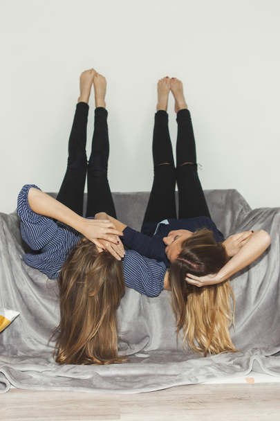 Two girls laying upside down on a grey sofa with legs up - Foto, Imagen