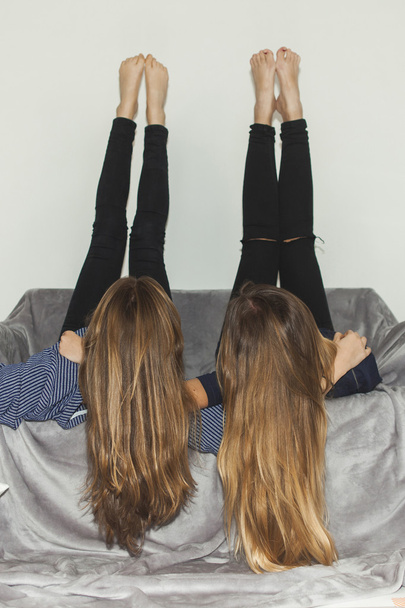 Two girls laying upside down on a grey sofa with legs up - Fotoğraf, Görsel