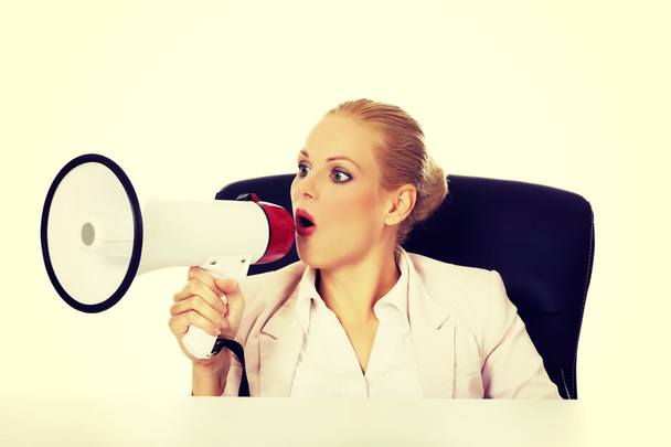 Business woman sitting behind the desk and screaming through a megaphone - Fotoğraf, Görsel