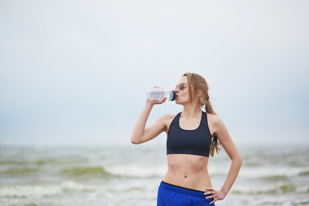 junge Fitnessläuferin trinkt Wasser am Strand - Foto, Bild