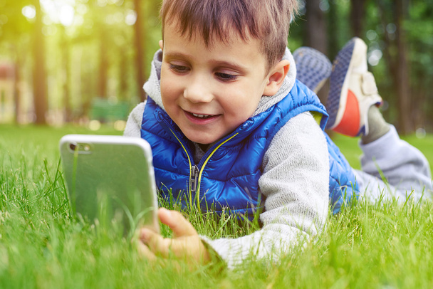  Μικρό αγόρι με έξυπνο τηλέφωνο που αναπαύεται σε πράσινο γρασίδι στο πάρκο - Φωτογραφία, εικόνα