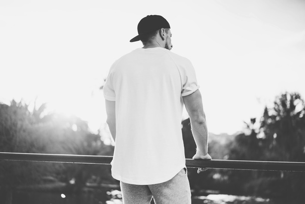 Photo Bearded Muscular Man Wearing Blank t-shirt, snapback cap and shorts in summer time. Green City Garden Park Sunset Background. Back view. Horizontal Mockup.Black White - Photo, image