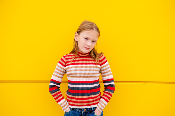 Portrait of a cute little girl of 8 years old, wearing stripe roll neck pullover and denim skirt, standing next to bright yellow wall - Valokuva, kuva