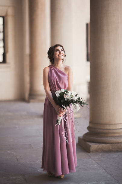 Portrait of a beautiful young bride that stands between the columns near old building and holding bridal bouquet - Photo, image