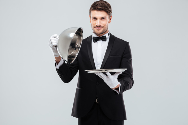 Handsome young butler in tuxedo opening metal tray with lid - Foto, immagini