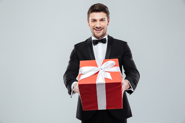 Cheerful attractive man in tuxedo giving you red gift box - Фото, изображение