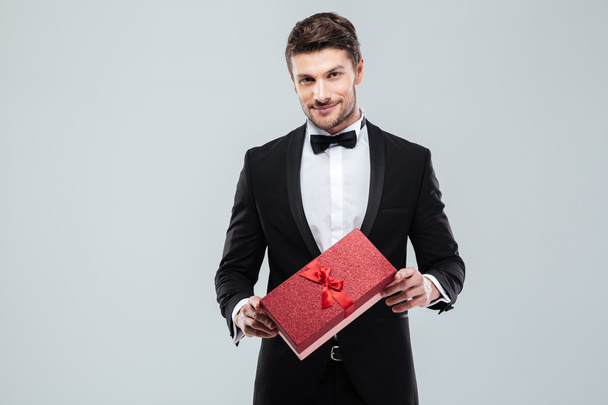 Confident attracive man in tuxedo standing and holding gift box - Fotó, kép