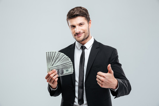 Smiling young businessman holding money and showing thumbs up - Foto, Bild
