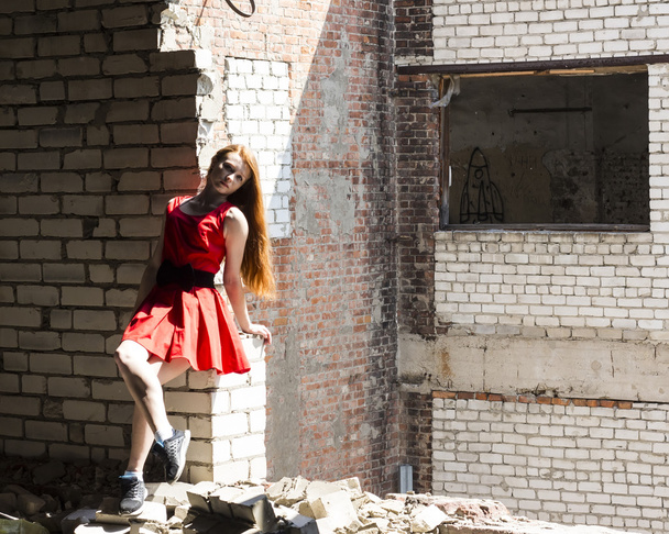 Red-haired thin slim girl with freckles  in red dress on  background of  wall of  destroyed building - Zdjęcie, obraz