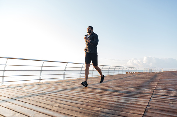 Sportsman listening to music and running on pier at sunset - Photo, Image