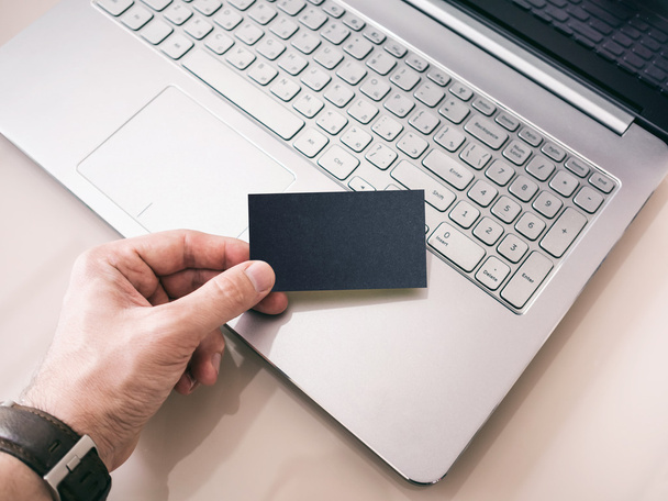 Man's hand holding blank Black Business Card. Modern Laptop on background, mockup - Zdjęcie, obraz