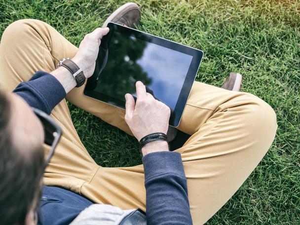 Man using a digital Tablet Mockup with blank screen sitting on the grass in the park - Фото, изображение
