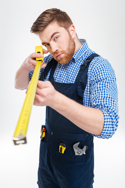Concentrated bearded young worker in overalls using measuring tape - Valokuva, kuva