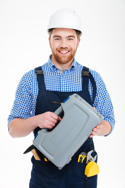 Smiling bearded young worker in helmet holding tool box - Fotó, kép