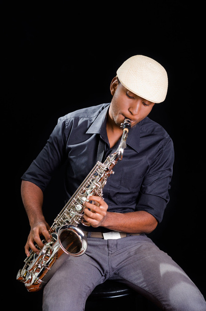 With white hat a black man is sitting and playing his saxophone, dark background, nice music - Foto, imagen