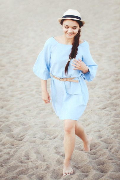 Portrait of smiling laughing white Caucasian brunette woman with tanned skin in blue dress and straw hat running on sand beach, sunset on summer day, lifestyle concept - Foto, afbeelding
