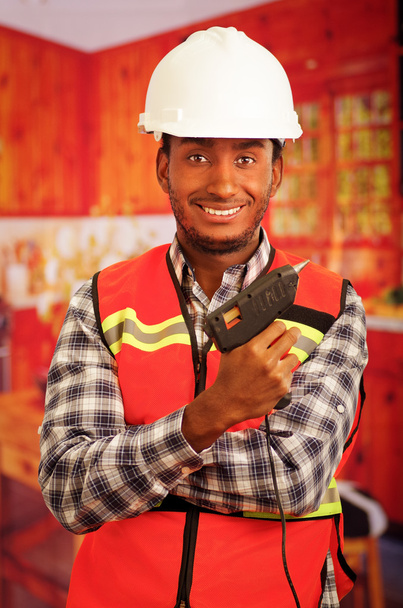 Young engineer carpenter wearing square pattern flanel shirt with red safety vest, holding glue gun smiling to camera - Fotografie, Obrázek