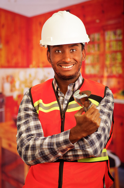 Young engineer carpenter wearing helmet, square pattern flanel shirt with red safety vest, holding small handheld electric saw smiling to camera - Foto, Imagen