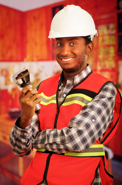 Young engineer carpenter wearing helmet, square pattern flanel shirt with red safety vest, holding small handheld electric saw smiling to camera - Foto, imagen